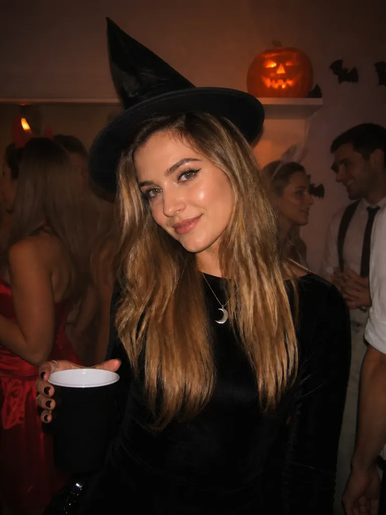 A woman in a witch hat holding a cup at a Halloween house party, a lit jack-o'-lantern on the shelf behind her