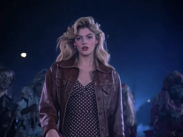 The lead in a brown leather jacket walks toward the camera, zombies close behind her on a night street