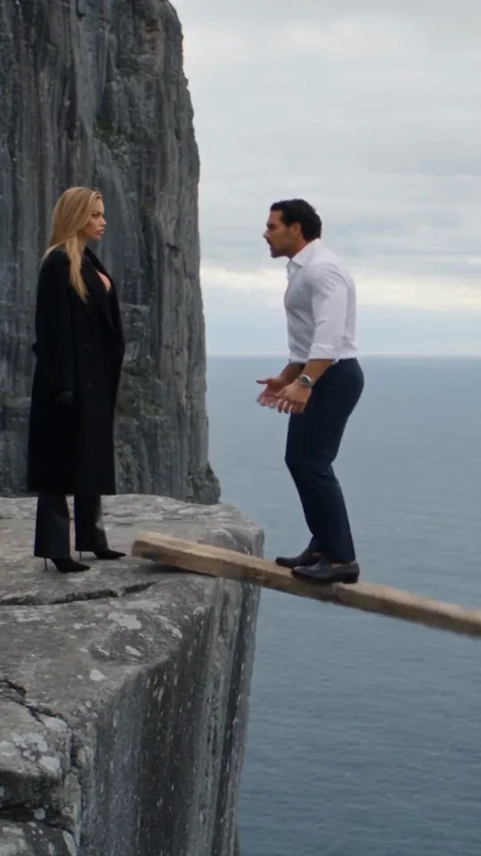 Wide shot: the woman in the long black coat stands back on the cliff edge as the plank tips up under the man in the white shirt