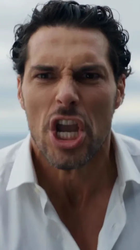Close-up of a dark-haired man in an open white shirt yelling, mouth wide open, against a grey sky