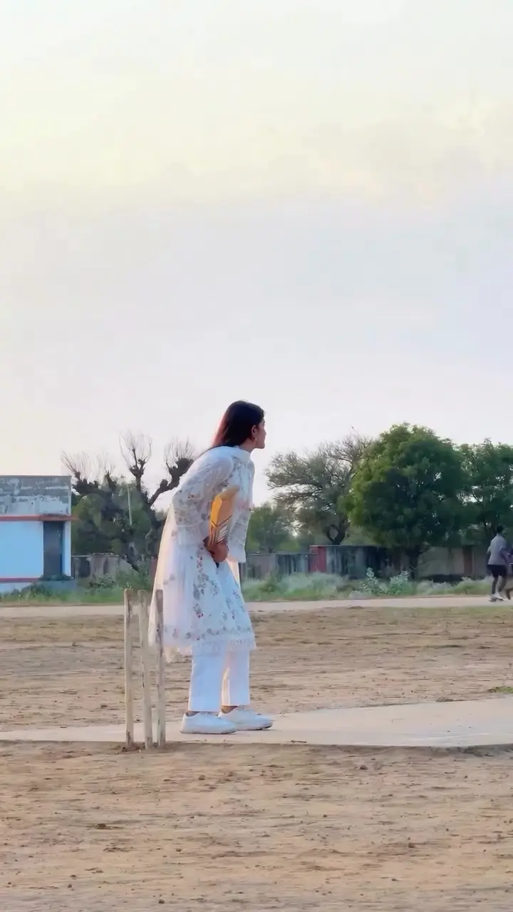 A batter stands side-on at the stumps with the bat low, a pale building behind