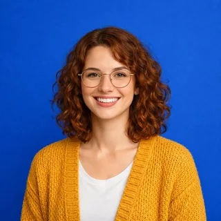 A smiling woman with curly auburn hair and round glasses, alone against a blue backdrop