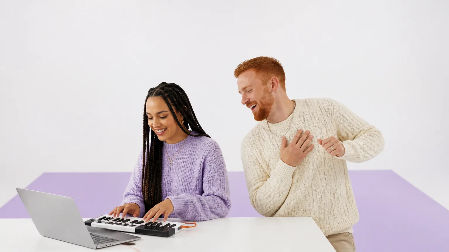 A couple in a white studio, one playing a keyboard and the other leaning in mid-dance, laughing
