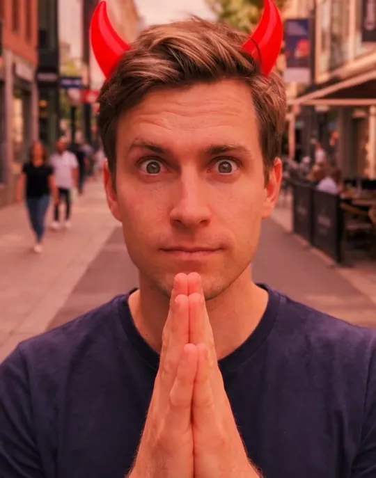 A man on a city street keeps his palms pressed together, wide-eyed, as red light fills the frame and two red horns grow from his hair