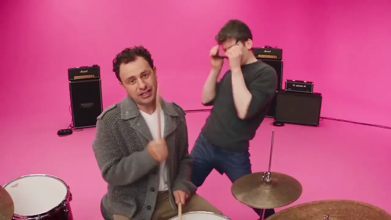 Two men in a hot-pink studio, one playing the drums and one dancing behind him