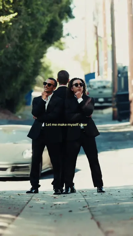 Three dancers in black suits posing in a tight huddle on a sunny pavement, the middle one turned away from the camera