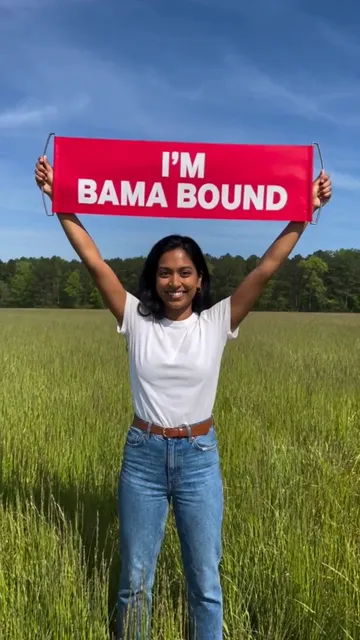 穿白 T 恤和牛仔裤的年轻女生在绿色草地上把红色 I'M BAMA BOUND 横幅举过头顶，对着镜头微笑