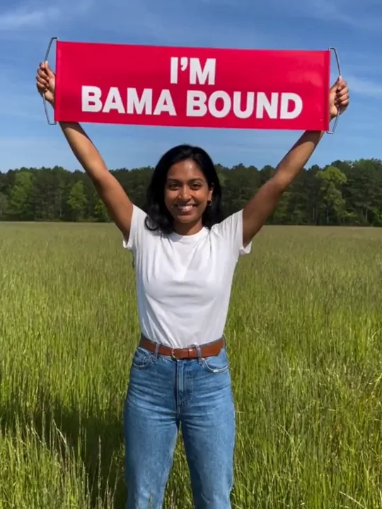 穿白 T 恤和牛仔裤的年轻女生在绿色草地上把 I'M BAMA BOUND 横幅举过头顶。