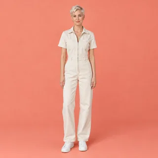 A woman with a platinum pixie cut in a cream jumpsuit standing full-length against a coral backdrop