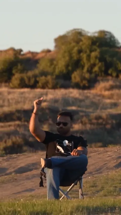 A man in sunglasses and a black T-shirt sits cross-legged in a camp chair in a dry field, one hand raised to snap