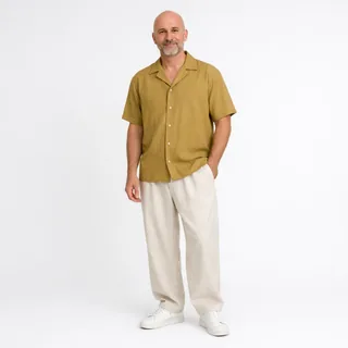 A bald man with a grey beard in a mustard short-sleeve shirt, cream linen trousers and white sneakers stands full length on a white background.