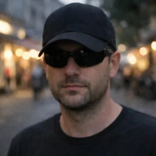 A man in dark sunglasses and a low cap, his face in shadow, on a blurred evening street.