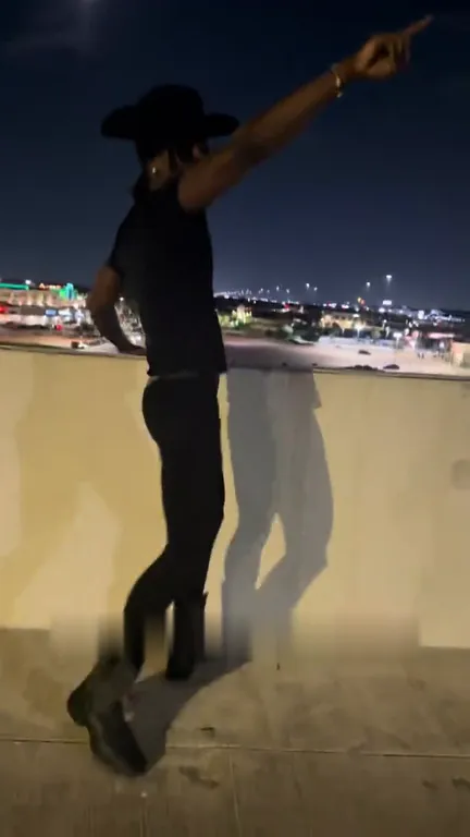 Singer in a black cowboy hat pointing at the night sky with one leg kicked out on a rooftop