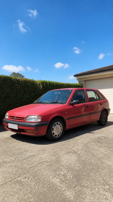A faded red hatchback parked on a sunny driveway in front of a hedge and a garage