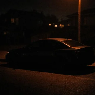 A dark sedan under a dim streetlight at night, mostly lost in shadow