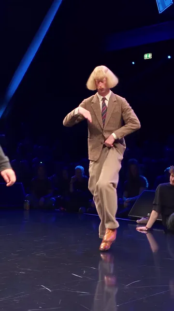 Dancer in a tweed suit and blonde bob lifting one knee, forearms folded at his chest