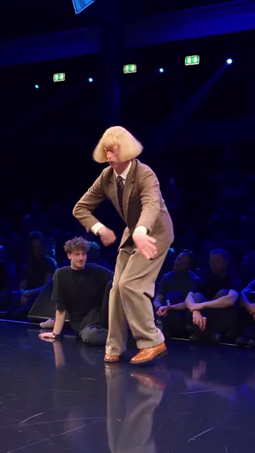 Dancer bent low in the suit, arms dangling, a row of seated spectators behind him
