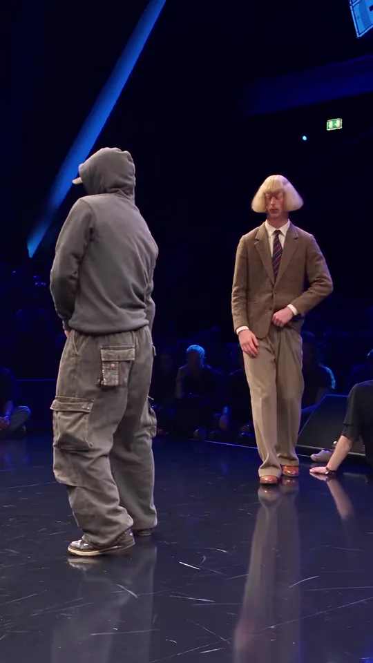 A hooded opponent in grey faces a dancer in a brown tweed suit and blonde bob on a dark battle stage, the crowd seated behind them