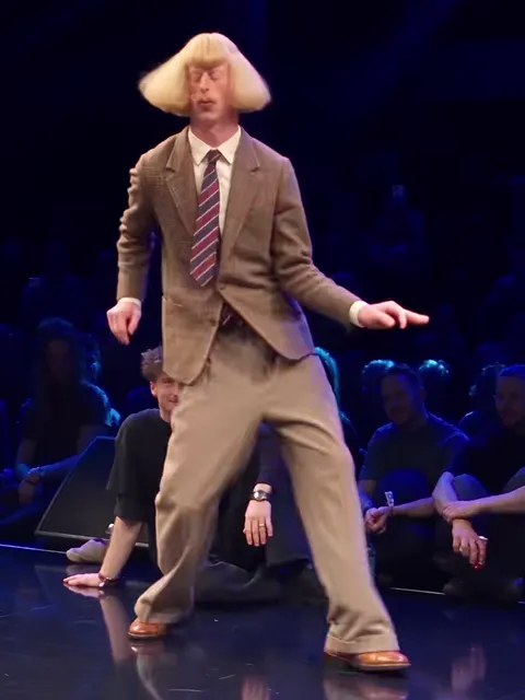 The dancer in a brown tweed suit, striped tie and wide beige trousers, in a wide stance on the stage