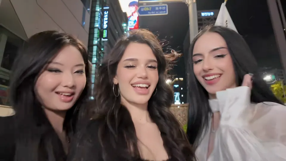 三位朋友在首尔夜街挤在一起自拍，身后是高楼和亮着的招牌
