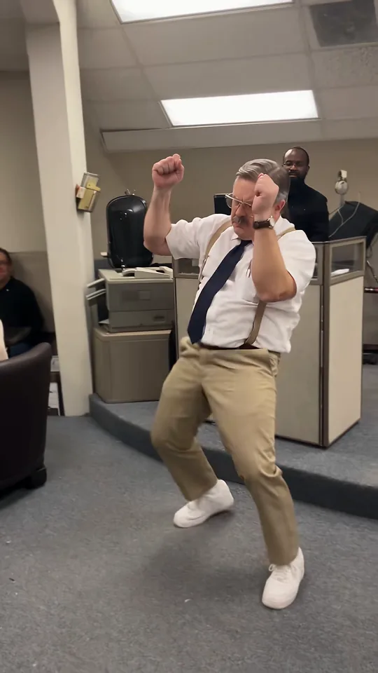 The dancer with both fists beside his ears, elbows out, mid knee-lift in the office
