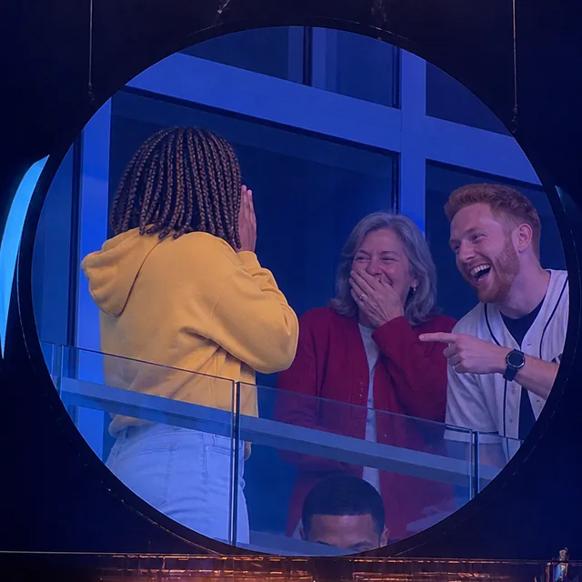 Kiss cam AI jumbotron meme: the person in front stands with their back to the camera while the person behind has ducked below the railing and fans in the next box laugh