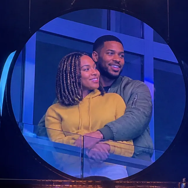 Two people hugging from behind in a stadium box, shown on a round kiss cam screen under the lighting rig