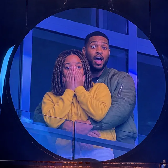 On the kiss camera big screen, the person in front covers their face with both hands while the person behind stares at the lens