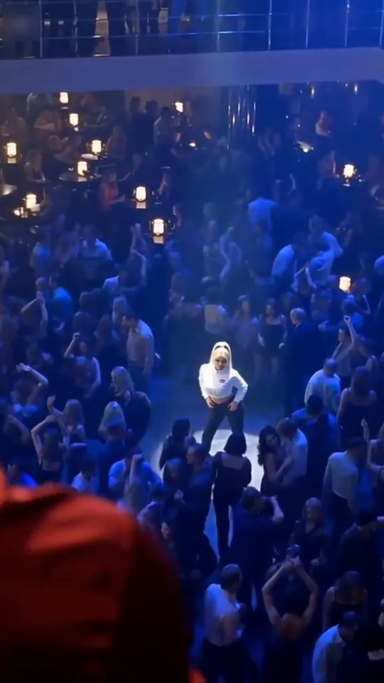 View over the watcher's shoulder down to a crowded blue dance floor with one person in a white spotlight