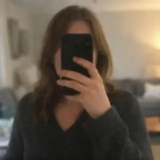 A woman takes a mirror selfie with her phone covering most of her face, a blurred living room behind