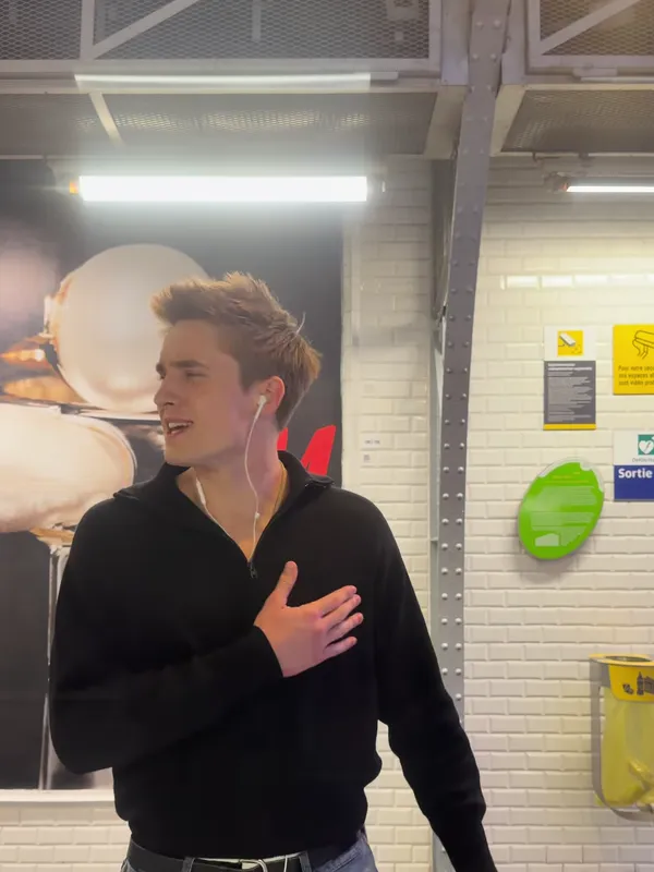 A young man in a black zip sweater singing with a hand on his heart on a Paris metro platform