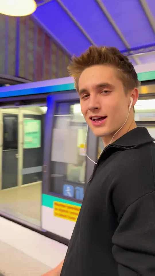Close-up of a young man in a black zip sweater smiling into the camera on a Paris metro platform, a green and white train behind him