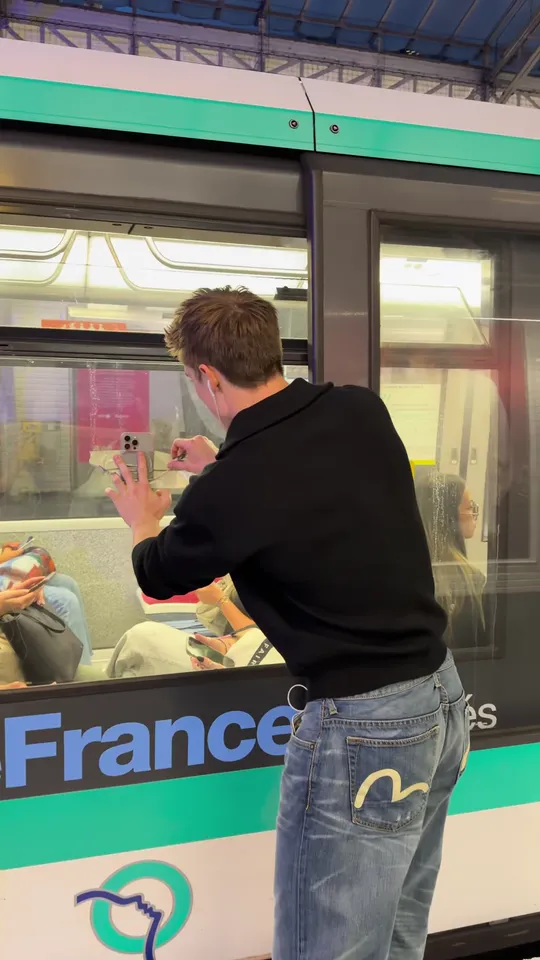 A young man seen from behind pressing a phone against the outside of a metro window while passengers sit inside