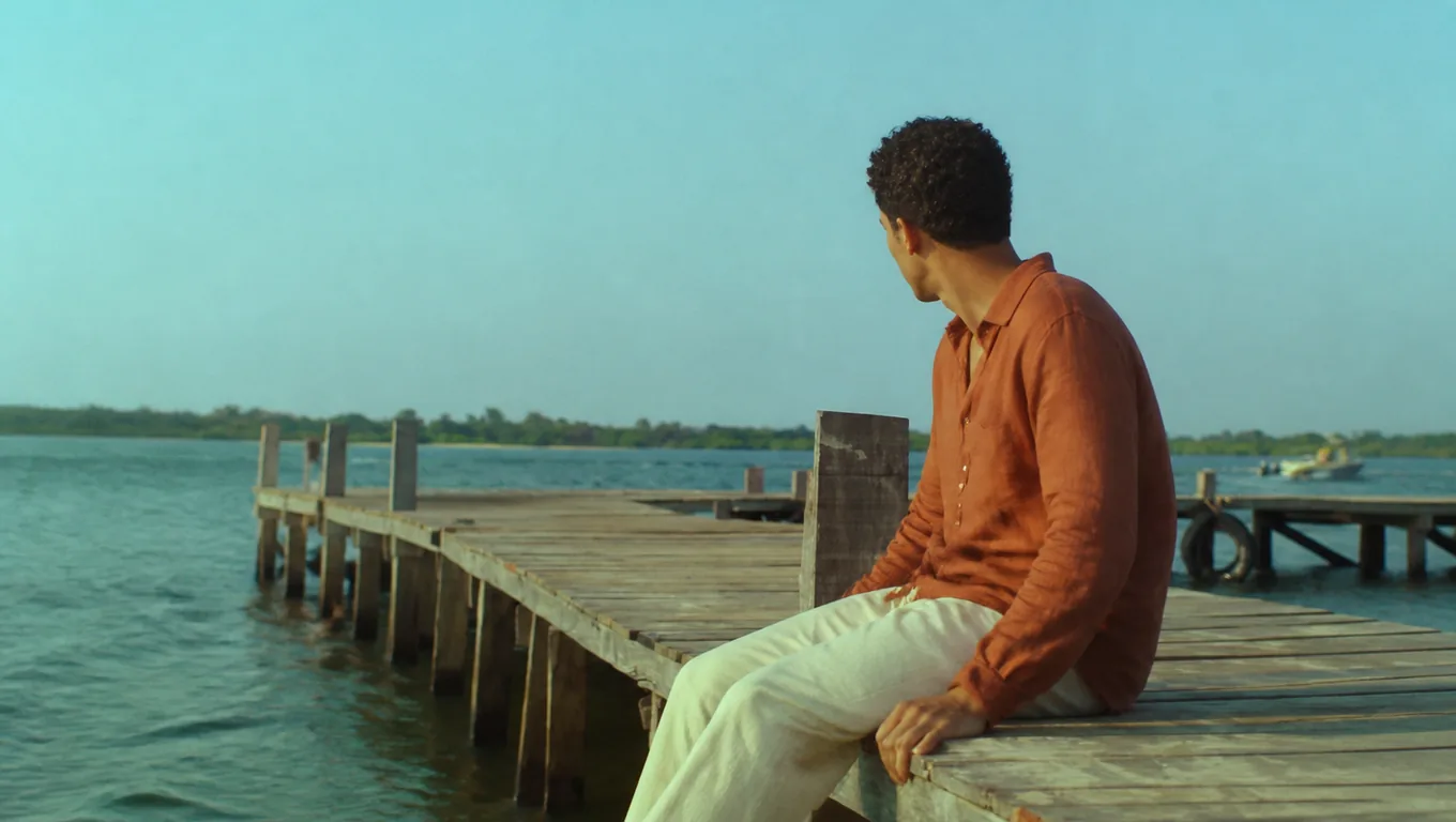 A man in a rust-orange linen shirt sits on the edge of a wooden pier and looks back over his shoulder