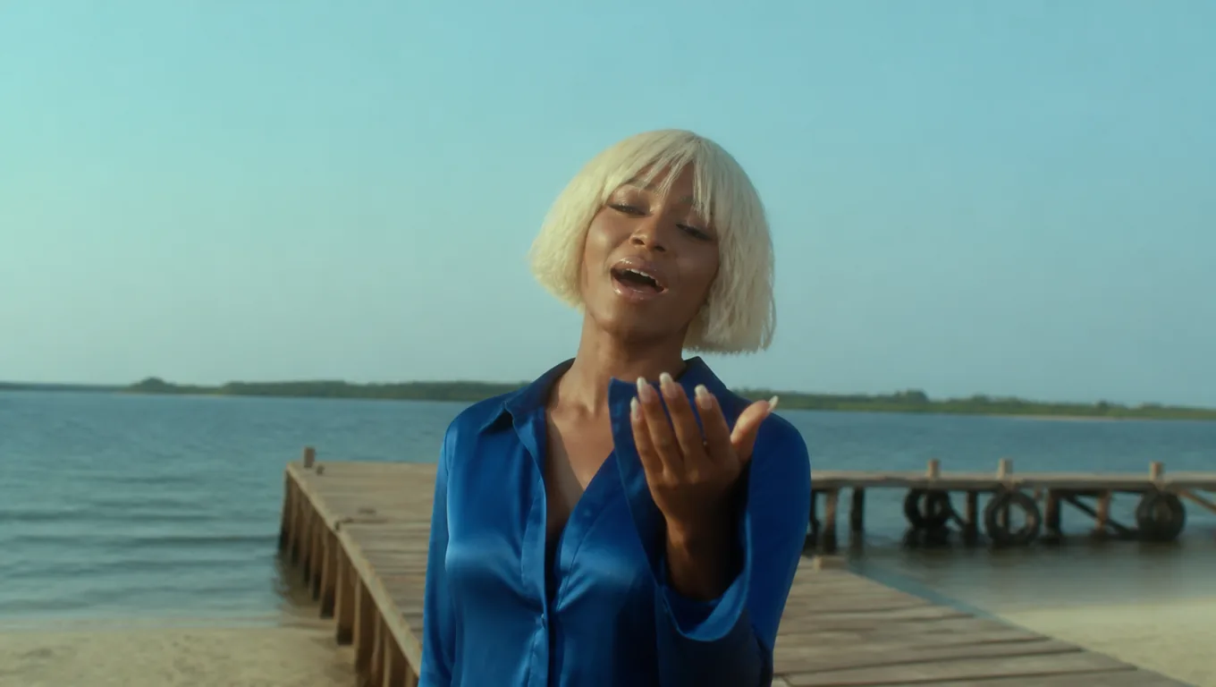 A woman with a platinum bob in a blue satin shirt sings toward the camera with one hand raised at the end of a wooden pier
