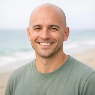 A smiling man in a sage T-shirt in bright daylight, the beach blurred behind him