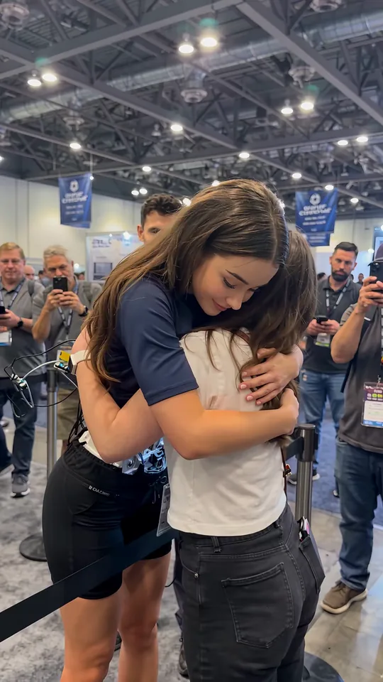 AI robot hug at a tech expo: the android rests her head on the visitor's shoulder, eyes closed and smiling, as people film