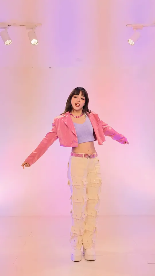 The dancer stands in a pink-lit studio with both arms swung out to the sides