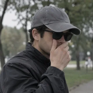 Man in dark sunglasses and a cap covering his mouth with one hand in a blurred park