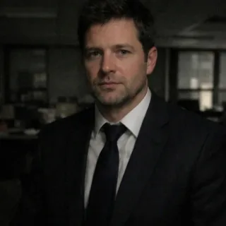 Man in a dark navy suit and tie that almost disappears into a dim, blurred office