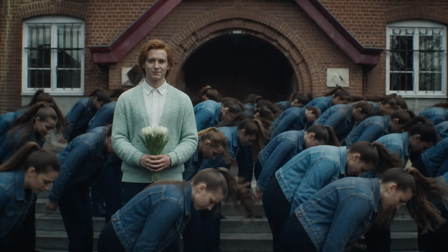 A red-haired young man in a mint cardigan holds white tulips and stands still on school steps as rows of the same young woman in a denim jacket whip forward around him