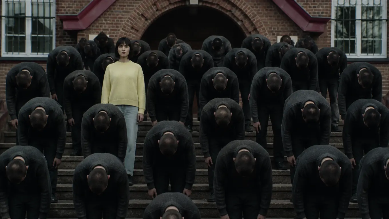 A woman in a pale yellow sweater stands upright on brick school steps while rows of identical men in charcoal sweaters bow forward around her