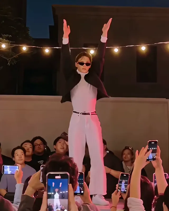 The dancer in sunglasses and a black jacket raising both arms straight up overhead