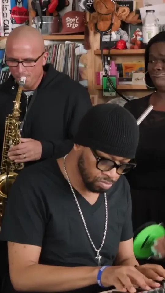 Keyboard player in a black beanie and glasses at the front, bald sax player in a black hoodie behind, bookshelves full of souvenirs