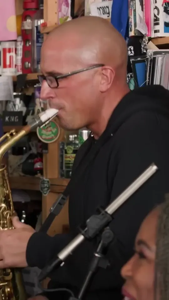 Bald sax player in glasses playing a solo side-on, a mic stand in front and a smiling backing singer below
