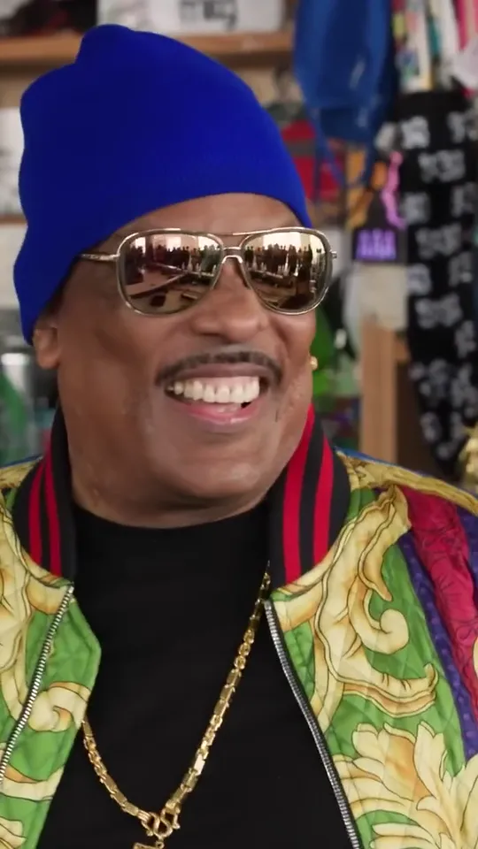 Singer in a blue beanie, gold mirrored sunglasses and a patterned jacket grinning at the camera