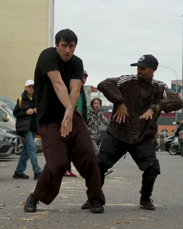 Both dancers swing their arms down across their bodies while two people walk past behind them