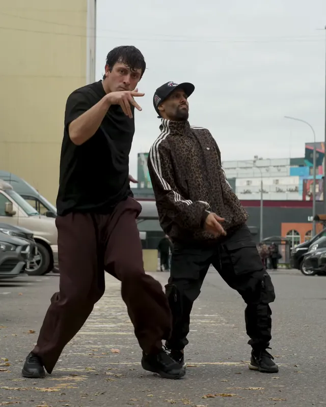 The two dancers lean together shoulder to shoulder, the one in the black tee pointing at the camera