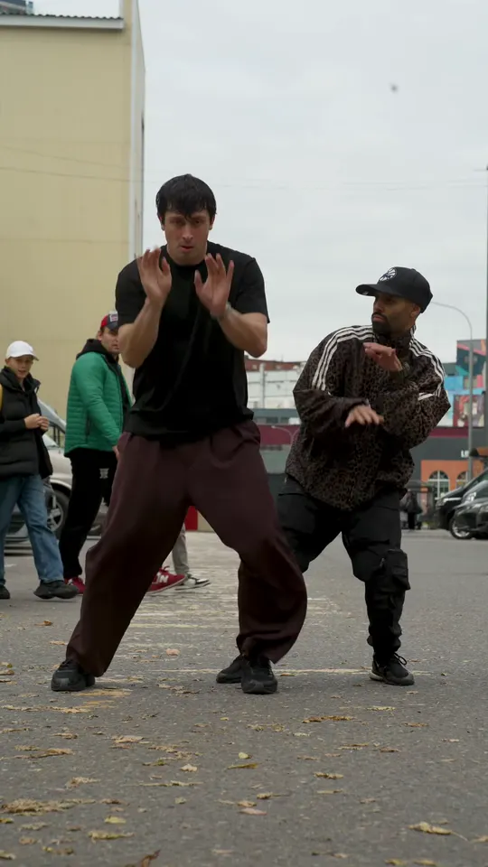 Mid-routine in the grey parking lot: the dancer in the black tee holds both hands up, the dancer in the cap squats low beside him, and people walk past behind them