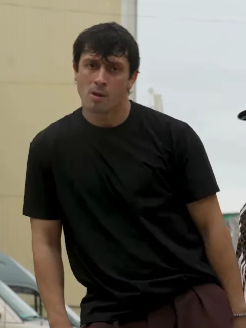The left dancer: dark hair and a black T-shirt, waist up in the grey parking lot